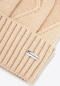 Cappello beige lavorato a maglia con texture a coste e motivo intrecciato. Presenta una targhetta argentata con "WITTCHEN" sul lato.