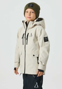 Beige wasserdichte Jacke mit Kapuze, schwarzem Reißverschluss, vier Taschen und Logodetails. Kombiniert mit schwarzer Hose und grüner Strickmütze.