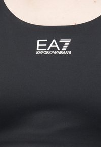 Fekete testhez simuló trikó, sima textúrával és középen a mellkason egy hangsúlyos fehér logóval, amelyen az "EA7 EMPORIO ARMANI" felirat olvasható.