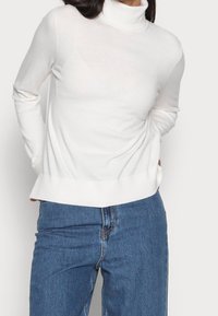 Pull à col roulé blanc à manches longues, texture douce et coupe décontractée, associé à un jean en denim bleu.