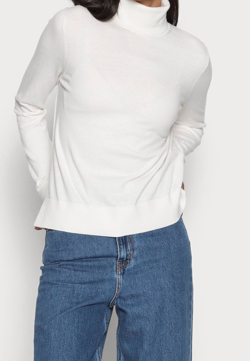 Pull à col roulé blanc à manches longues, texture douce et coupe décontractée, associé à un jean en denim bleu.