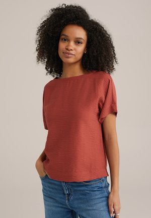 Rust-colored short-sleeve top with a textured fabric, boat neckline, and slightly rolled sleeves, paired with blue denim jeans.