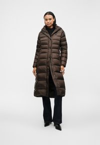 Brun, lang pufferjakke med hætte, quiltet design og frontknapper. Båret over mørke, flagrende jeans og spidse sorte hæle. Glat tekstur.
