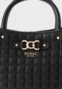 Juodas siuvinėtas odinis Guess rankinis krepšys su dviem rankenomis, auksinės spalvos detalėmis ir "GUESS EST 1981" logotipu priekyje.