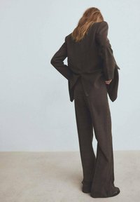 Osoba z długimi włosami, ubrana w ciemnobrązowy, oversize'owy garnitur z szerokimi spodniami, stoi z rękami za plecami, opierając się o jasną ścianę.