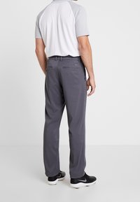 Man i grå golfbyxor, vit och grå kortärmad skjorta samt svarta golfskor som står vänd bortåt mot vit bakgrund.