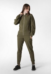 Giacca a zip verde oliva con un finish testurizzato e polsini elasticizzati, abbinata a pantaloni jogger coordinati e sneakers nere.