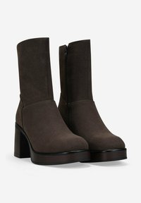 Botas de tobillo de ante marrón oscuro con tacones altos gruesos, punteras redondeadas y cremalleras laterales, sobre un fondo claro y liso.