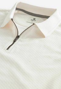 Hvid polo shirt med et struktureret mønster, lynlås lukning og et ternet kraveinteriør. Mærket siger "NEXT" med størrelse "M."