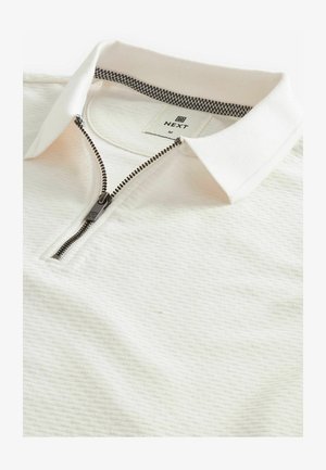 Hvid polo shirt med et struktureret mønster, lynlås lukning og et ternet kraveinteriør. Mærket siger "NEXT" med størrelse "M."