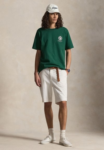 Junger Mann mit lockigem Haar, der eine weiße Ralph-Kappe, ein grünes kurzärmliges Hemd, weiße Shorts, einen braunen geflochtenen Gürtel, weiße Socken und Turnschuhe trägt.