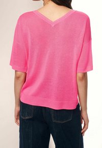Suéter de punto rosa con escote en V y mangas cortas, con dobladillo acanalado y un ajuste relajado, combinado con jeans de color azul oscuro. La textura parece ligera.