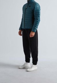 Teal puffjacka med horisontell sömnad, hög krage och fickor med dragkedja, kombinerad med svarta byxor i sweatpant-stil och vita sneakers.