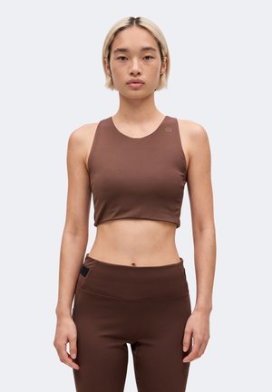 Noor isik lühikeste blondide juustega, kes kannab varrukateta pruuni lühikest topi ja vastavaid kõrge vöökohaga pükse lihtsa tausta ees.