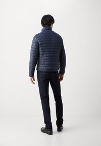 Marineblaue Steppjacke mit gesteppter Textur, hohem Kragen und figurbetontem Design, kombiniert mit dunklen Jeans und schwarzen Schuhen.