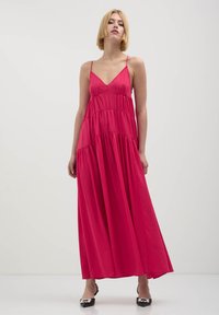 Donna con capelli biondi corti che indossa un vestito maxi a strati senza maniche di un vivace colore rosa e tacchi neri a punta con decorazioni in perle.