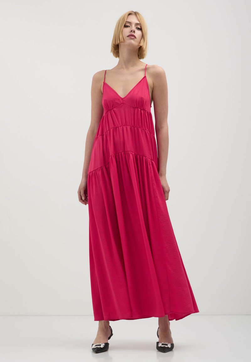 Donna con capelli biondi corti che indossa un vestito maxi a strati senza maniche di un vivace colore rosa e tacchi neri a punta con decorazioni in perle.