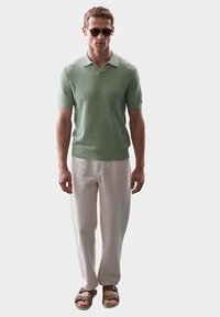 Lichtgroene gebreide polo met korte mouwen en een V-hals, gecombineerd met crèmekleurige broek en bruine sandalen. Gladde textuur, casual pasvorm.