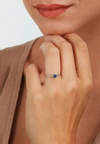 Sølvring med en rund blå ædelsten, sat på en slank ring, båret på en hånd som hviler nær hagen. Glat, poleret overflade.