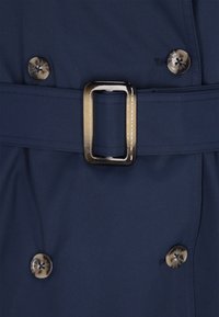 Marineblauer Trenchcoat-Stoff mit einem glänzenden Schnallenriemen und großen, runden braunen Knöpfen mit Kreuzmuster. Glatte Textur, maßgeschneiderte Passformdetails.