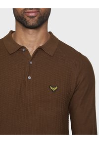 Braunes, strukturiertes Langarm-Polohemd mit Kragen, drei Knöpfen und einem gelben Emblem auf der linken Brust.
