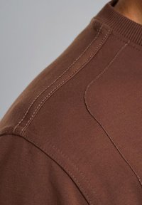 Sweatshirt en coton marron avec col ribbé, présentant une texture lisse et des coutures contrastées le long de la couture de l'épaule.