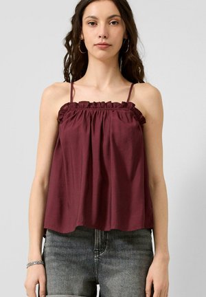 Jonge vrouw die een mouwloze bordeauxrode top met ruches en dunne bandjes draagt en vervaagde zwarte jeans met een hoge taille, staande tegen een effen achtergrond.