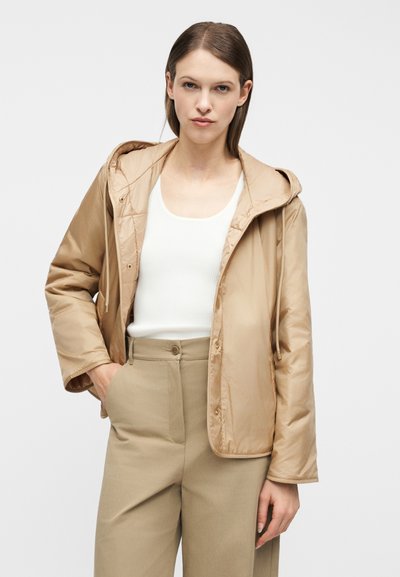 Νεαρή γυναίκα που φοράει μπεζ σακάκι με κουκούλα, λευκό τοπάκι και μπεζ παντελόνι ψηλόμεσο, στέκεται με το ένα χέρι στην τσέπη μπροστά σε απλό φόντο.