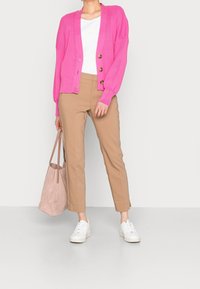 Cardigan rosa a coste con tre bottoni, abbinato a pantaloni beige e sneakers bianche. Il modello tiene una borsa a spalla rosa chiaro.