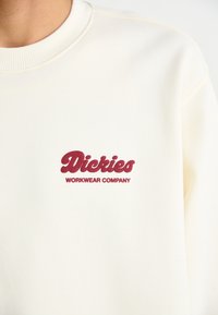 Cream sweatshirt featuring a ribbed neckline and a burgundy embroidered logo that reads "Dickies WORKWEAR COMPANY" on the chest.
