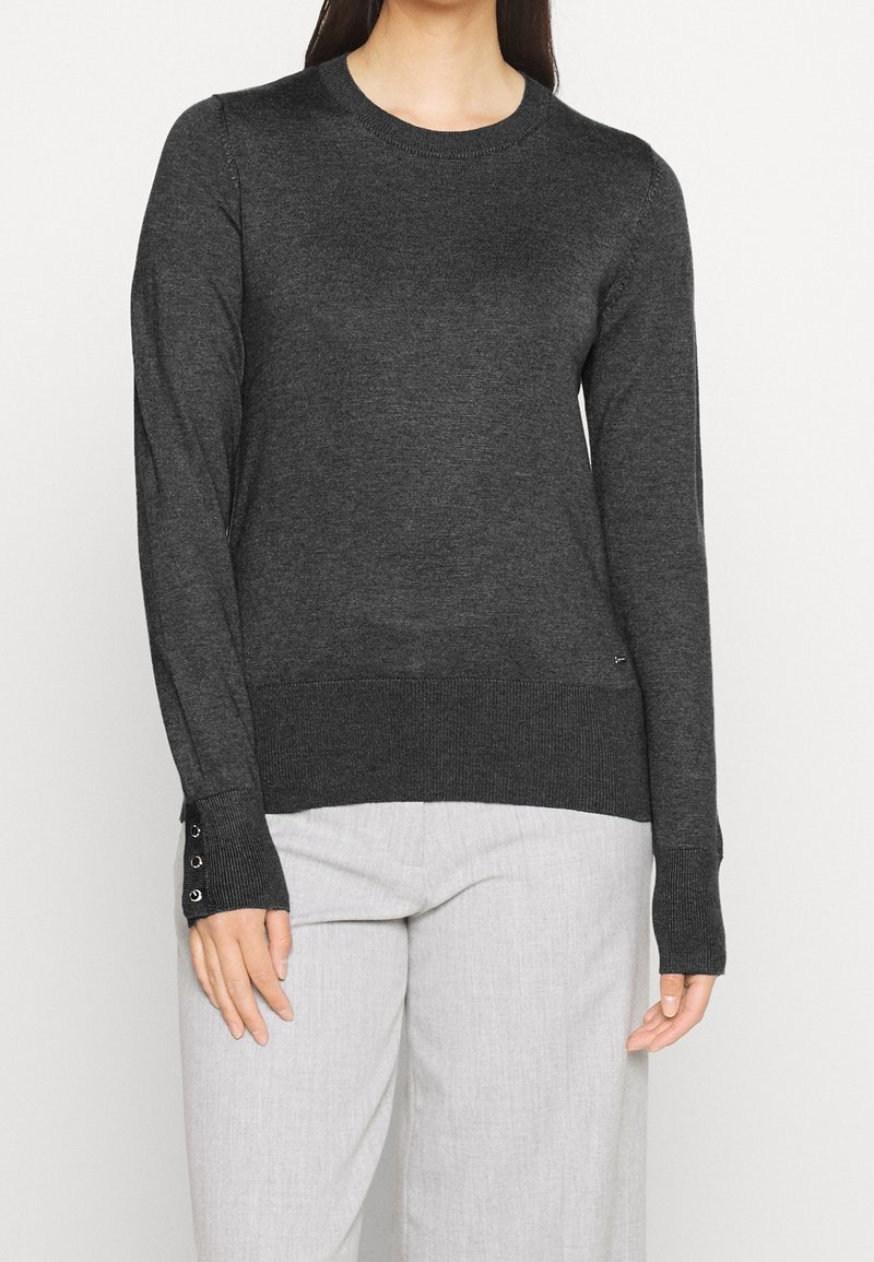Pull à manches longues gris foncé avec col rond et détails de boutons aux poignets, porté avec un pantalon gris clair, vue de face, visage non visible.