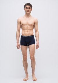 Νέος άνδρας όρθιος, ξυπόλυτος, φορώντας μπλε ναυτικό εσώρουχο Polo Ralph Lauren, σε απλό φωτεινό γκρι φόντο.