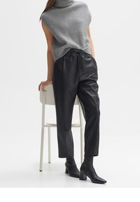 Czarne skórzane spodnie z elastycznym pasem, zestawione z szarym, prążkowanym topem bez rękawów. Modelka stoi obok białego krzesła.