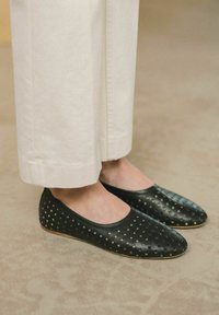 Bailarinas negras perforadas con un acabado suave y punta redonda. Presentan una suela flexible y se sitúan debajo de unos pantalones de pierna ancha blancos.