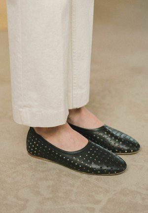 Ballerine nere perforate con finitura liscia e punta rotonda. Presentano una suola flessibile e si trovano sotto pantaloni bianchi a gamba larga.