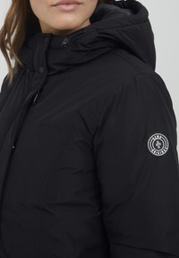 Svart dunjakke med hette, med et logo-patch på ermet, glatt tekstur og glidelås. Detaljert søm synlig.