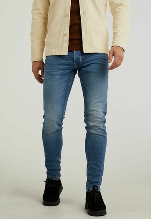 Homme portant un jean skinny bleu délavé, des chaussures noires, un pull marron et une veste beige en velours côtelé, debout devant un fond uni.