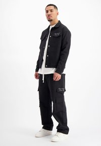 Zwarte denimjack met drukknoopsluiting, gecombineerd met zwarte cargobroek. Witte t-shirt eronder en witte sneakers. Geborduurde details op de outfit.