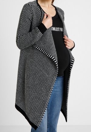 Osoba w ciąży w czarno-białym, wzorzystym otwartym kardiganie, czarnej koszulce z częściowym białym napisem oraz niebieskich dżinsach.