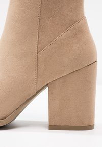 Bottine en daim beige avec un talon bloc et des coutures visibles, vue de profil sur fond blanc.