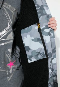 Vadderad jacka med svart fleece inuti och grått camouflagemönster på utsidan. Har en ficka med dragkedja i gult och grafisk design.