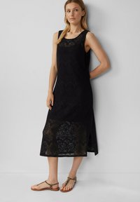 Zwart mouwloos lace jurk met intrikate patronen en een knielange snit, gecombineerd met bruine sandalen.