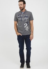 Grå piké med krave har trykte grafikker i blått og hvitt, kombinert med mørkeblå jeans og svarte sneakers.