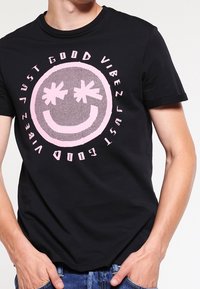 Musta puuvilla T-paita, jossa on suuri vaaleanpunainen hymiö tähtisilmin, ympärillä teksti "PELKKÄ HYVÄT VIBRAT."