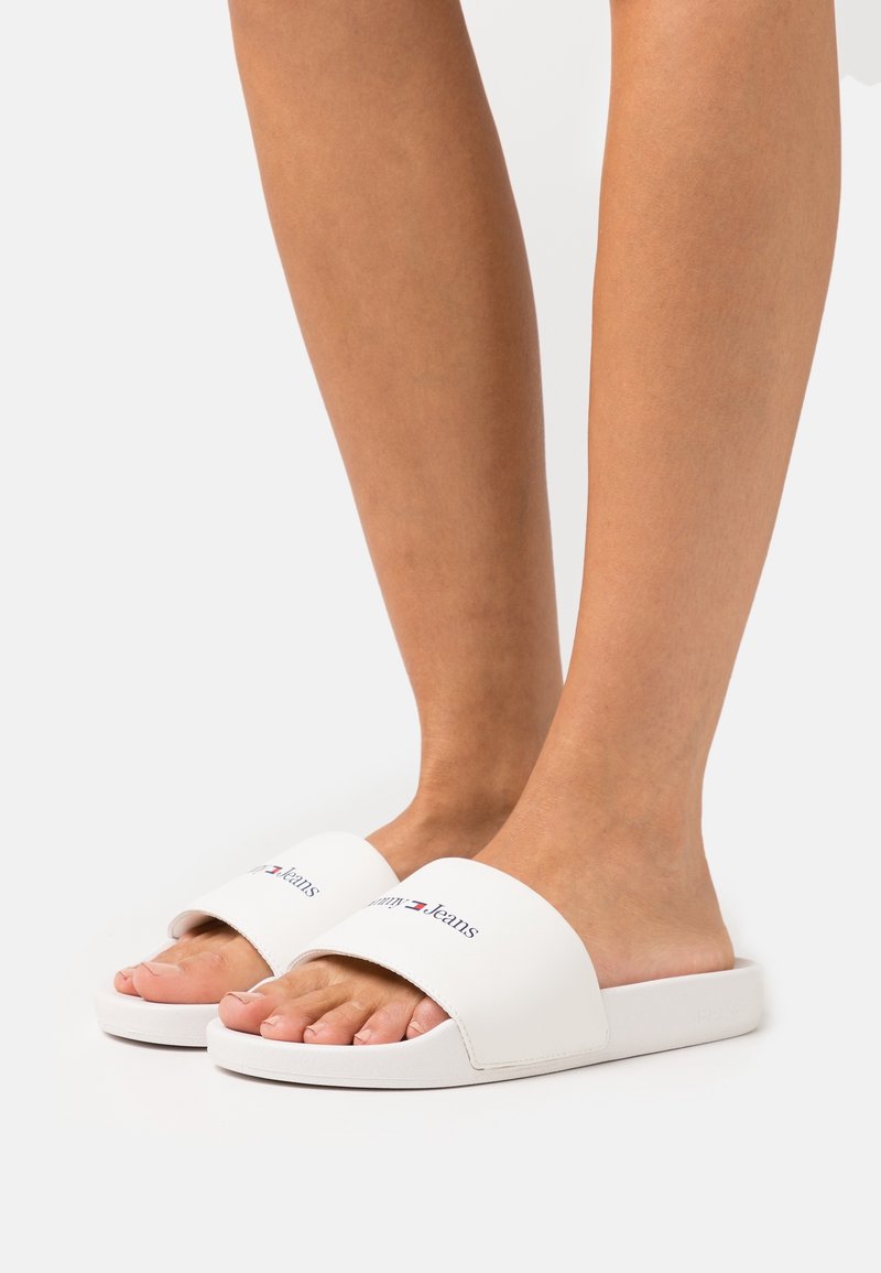 Pieds nus portant des sandales blanches à enfiler avec le logo "Tommy Jeans" sur la bride sur un fond clair uni.
