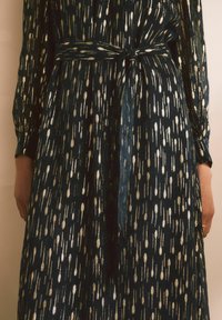 Long-sleeved dress in dark teal with a metallic gold drop pattern. Features a defined waist with a tied belt and a flowing silhouette.
