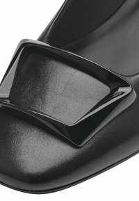 Svart sko med en slät lädertextur, prydd med en glänsande svart rektangulär spänneaccent på tån.