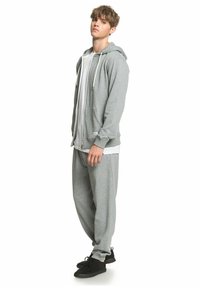 Sudadera con cremallera gris y cordones blancos, combinada con pantalones gris a juego. El tejido parece suave y texturizado, complementado por zapatos negros.
