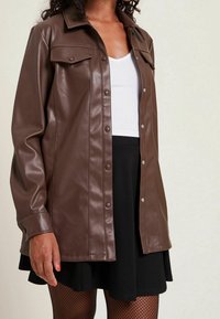 Giacca camicia in finta pelle marrone con bottoni anteriori, due tasche sul petto e una vestibilità leggermente rilassata. Abbinata a un top bianco e una gonna nera.