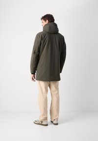 Olijfgroene waterdichte lange jas met een capuchon, met een rechte snit, elastische manchetten en een zachte textuur. Gecombineerd met lichtbeige broeken.